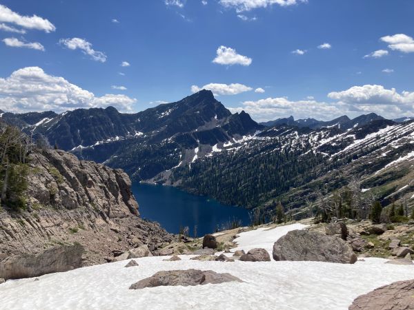 A scenic mountain landscape with a deep blue alpine lake, rugged snow-dusted peaks, rocky terrain, and a bright, partly cloudy sky.