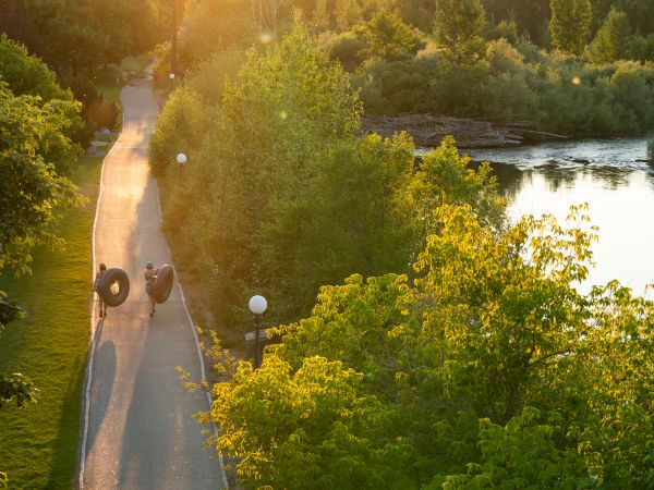 Image shows two people carrying tubes, walking on a sidewalk near the Clark Fork River in Missoula.