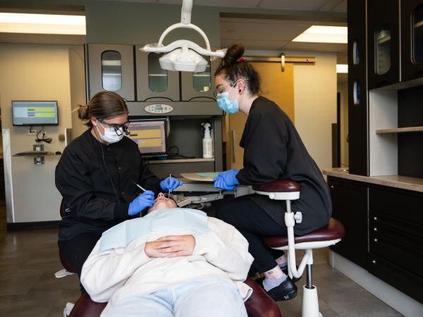 A student sits in a dental chair receiving care from a dentist and dental assistant. The dentist is examining the student’s teeth while the assistant stands nearby, preparing tools. The setting is a bright, clean dental clinic with equipment visible in the background.