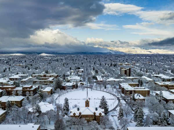 Aerial view of the 91��Ԫ campus in winter, with snow-covered buildings, trees, and walkways. The surrounding mountains are dusted with snow, and the Clark Fork River runs alongside the campus under a pale winter sky.