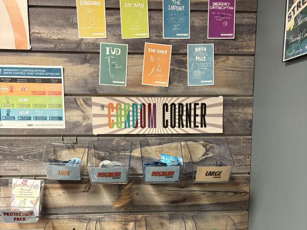 A wooden wall display titled “The Condom Corner” features educational posters about birth control methods and safer sex supplies. At the top are colorful informational cards describing options such as condoms, the ring, the implant, emergency contraception, IUDs, the shot, and birth control pills.  Below the posters are clear plastic bins mounted on the wall containing free safer sex supplies. The bins are labeled with different items including lube, regular condoms, large condoms, non-latex condoms, ultra-thin condoms, ribbed condoms, studded condoms, flavored condoms, internal condoms, and dental dams or finger condoms.  Additional educational posters about emergency contraception and sexual health are posted nearby, indicating this is a public health resource area providing information and free protection supplies.