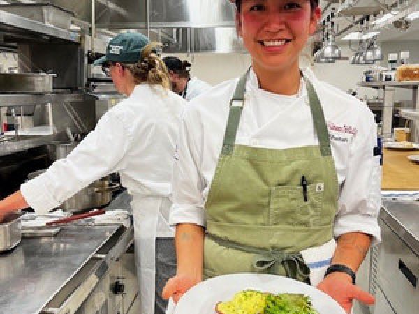 Chef in a commercial kitchen smiling while holding a plated dish topped with greens.