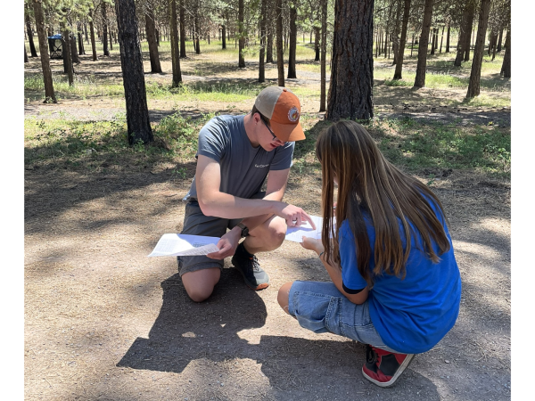 A graduate student and high school student kneel on the ground in Lubrecht Forest, comparing values for measurements.