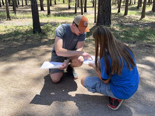 A role model kneels with a high school student  on the forest floor, comparing data on papers.
