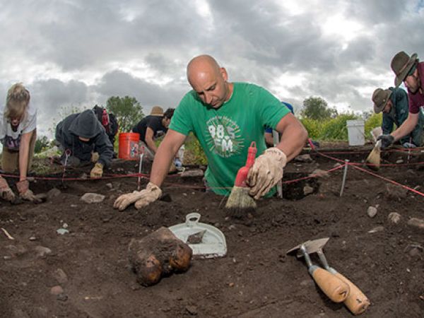 A man in a green shirt uses a brush to move dirt at an archaeological dig site. Other diggers are in the background. 
