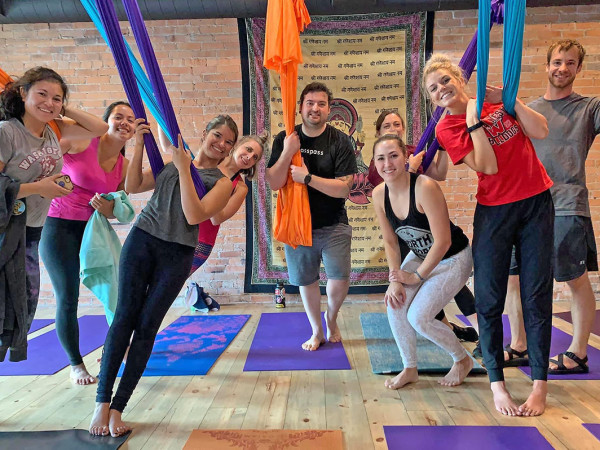 There are nine people in a yoga studio with colorful aerial yoga bands extending from the ceiling and yoga mats on the floor and a tapestry in the background 