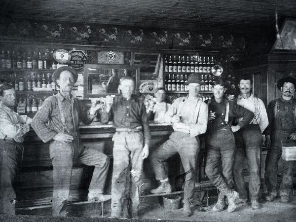 This is a very old photo of seven men standing in front of a saloon bar with bottles in the background 