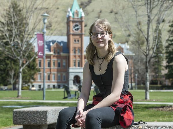 An image of a blonde-haired girl with glasses sitting on a bench with Main Hall blurred out in the background 