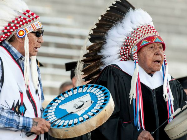 Two Native Americans, in eagle feather headdresses, sing and carry drums.