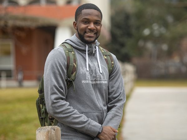 This is a portrait of a young Black man in a grey sweatshirt and a camo backpack smiling and a blurred out building in the background 