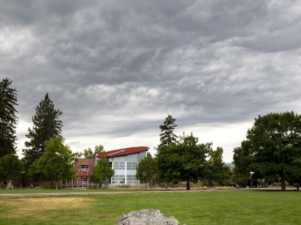 Student resting on boulder on oval with the PFNAC in the background standing out from the trees and grey clouds
