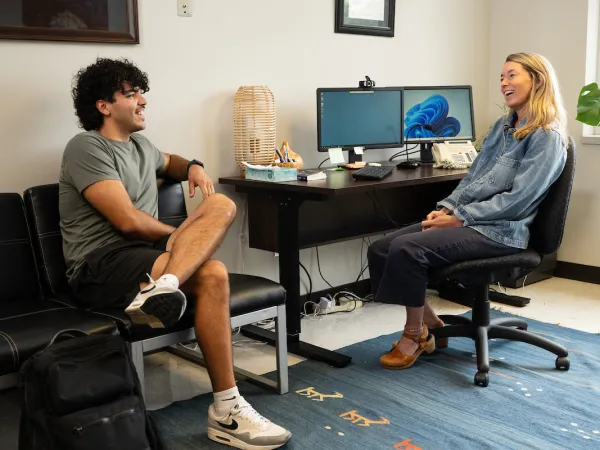 A man and woman sit smiling in an office. The man is on a couch with a backpack, and the woman is on a chair near a desk with monitors.