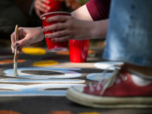 A University of Montana student in jeans paints colorful designs on pavement with a brush, holding a red cup. Red sneakers are visible nearby.
