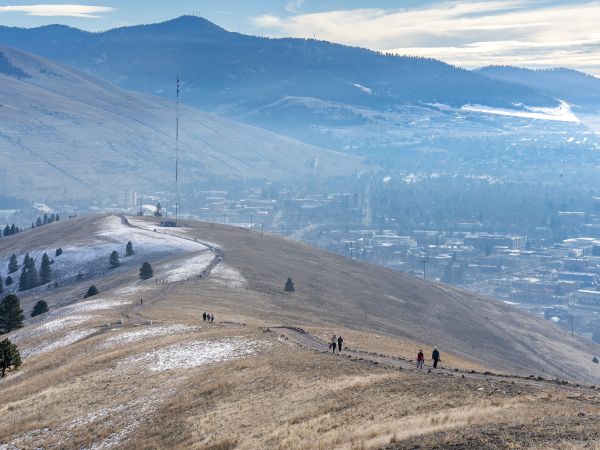 A wide view of a grassy hillside trail with patches of snow, where several people are hiking along a ridgeline above Missoula, with forested mountains and a hazy blue sky in the background.