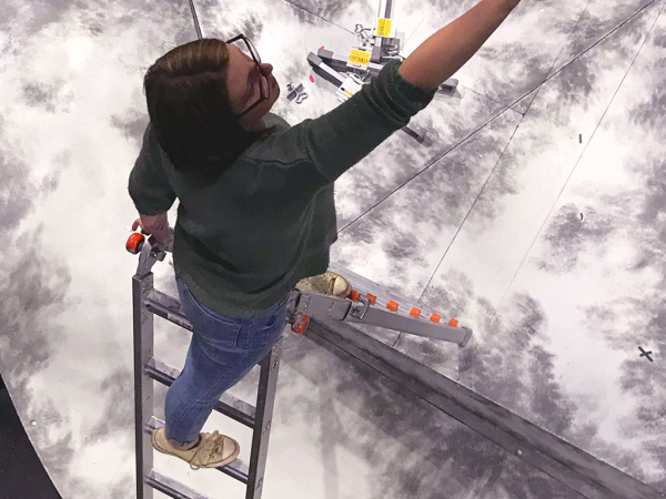 A design student paints a set atop a ladder, shot from above.