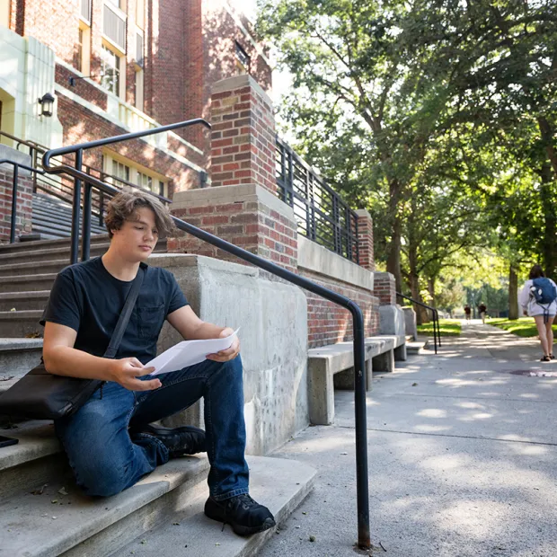 A student looks over a course syllabus while sitting on the steps of the Fine Arts Building