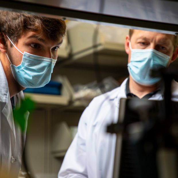 Two people wearing face masks and lab coats look at a computer monitor