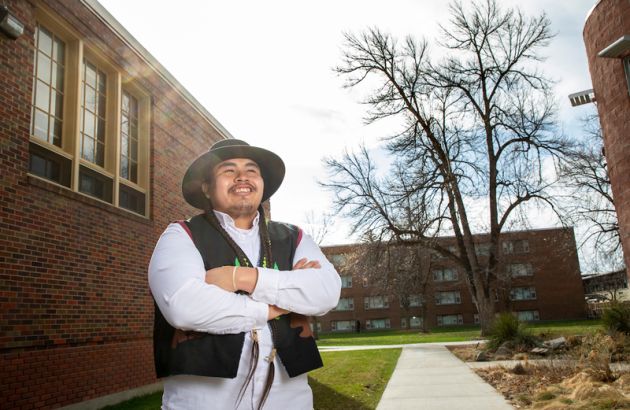 Zachariah Rides at the Door stands outside a brick building on campus. He wears a black hat and vest and holds his arms across his chest as he smiles.