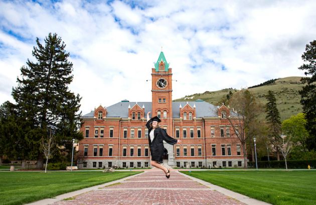 A graduate wears a cap and gown jumps in the air in front of Main Hall