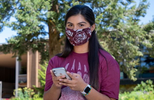 Female student wearing a cloth face covering holds her phone while standing in front of the UC