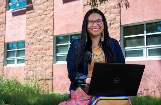 A student smiles at the camera while sitting outside the Payne Family Native American Center. There is a laptop on her lap.