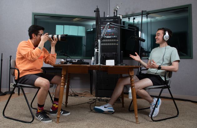 Two male students in audio booth, one taking a photo of the other talking into a microphone.