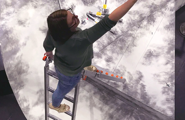 A design student paints a set atop a ladder, shot from above.