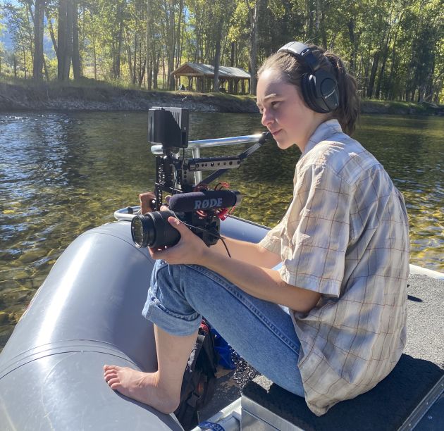 Woman with camera sits atop fishing boat on river.