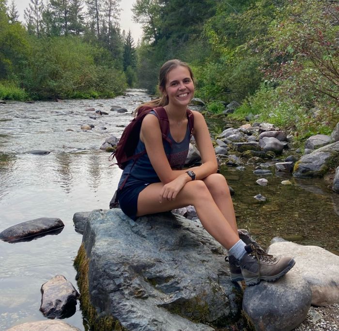 Audrey Glendenning smiles while sitting on a rock in a stream. She is wearing hiking clothes and boots and has backpack on.