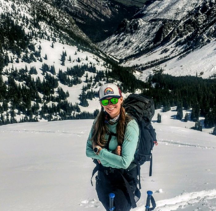 Kirsten Gerbatsch stands in a snowy field with her arms crossed as she smiles and looks at the camera. Two ski poles are visible in front of her.