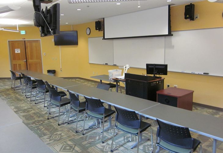 Classroom with U-shaped tables and chairs facing a projector screen, whiteboards, and an instructor podium.