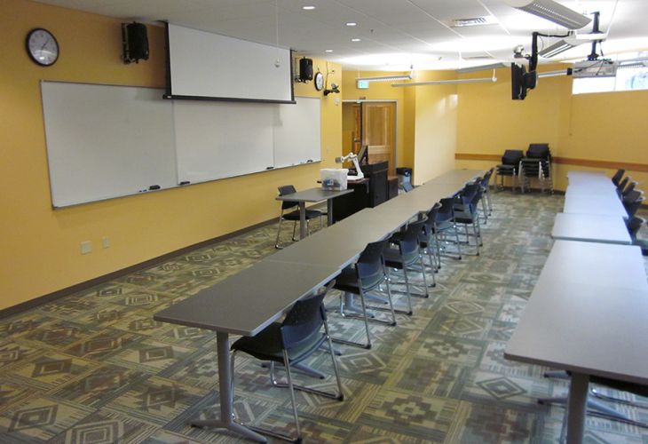 Empty classroom with long tables and chairs facing a projector screen and whiteboard.