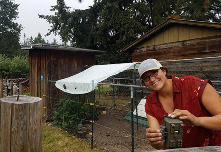A woman smiling while holding a wildlife camera in front of a fenced garden area with a wooden shed in the background.
