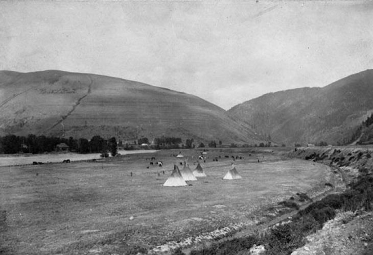 Historical black and white photograph of Native American tipis set against a mountainous landscape.