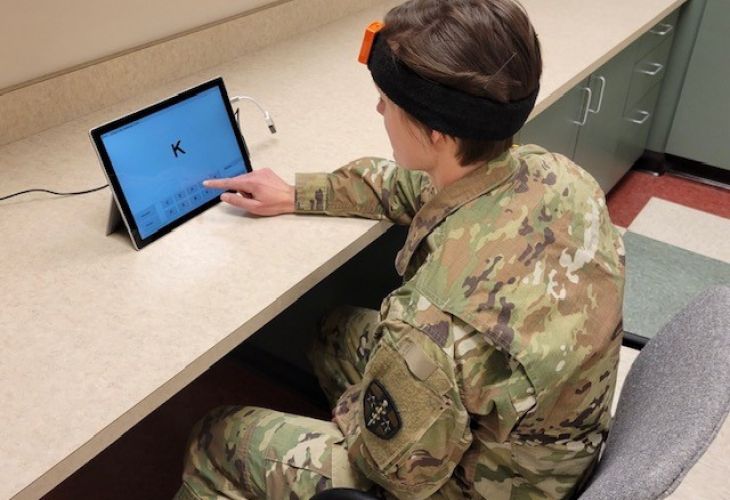 A soldier in camouflage uniform interacts with a touchscreen tablet while wearing a headband device.