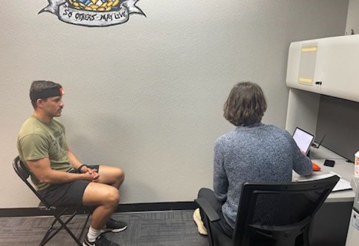 A man sitting in a chair with a headband while another person works on a laptop at a desk in an office setting.