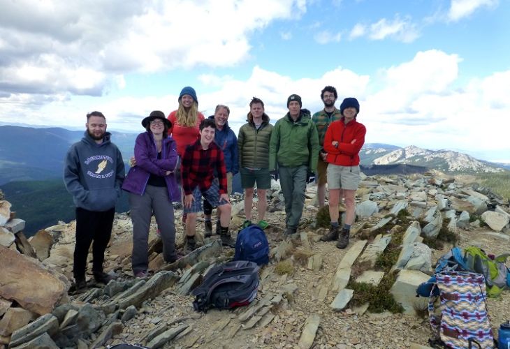 A group of nine hikers posing on a rocky mountain summit, with scenic views in the background.