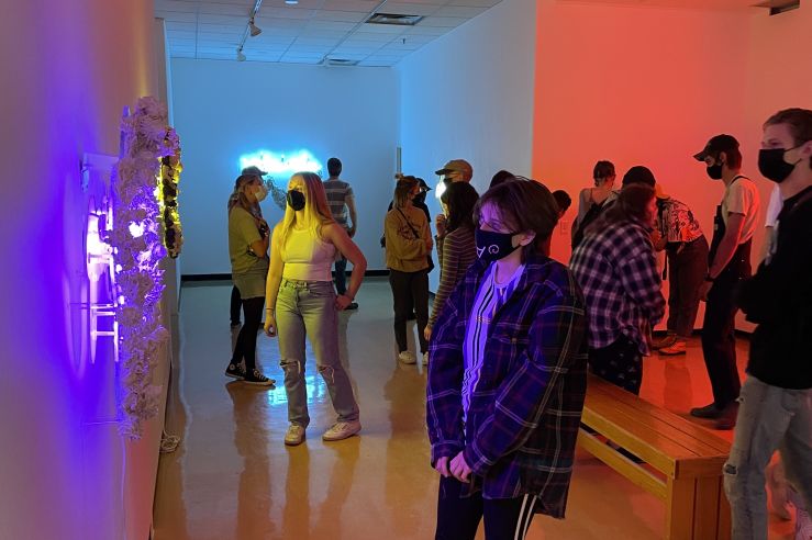 view of the gallery lit with purple, teal, and red neon lights with a light crowd of people in attendance