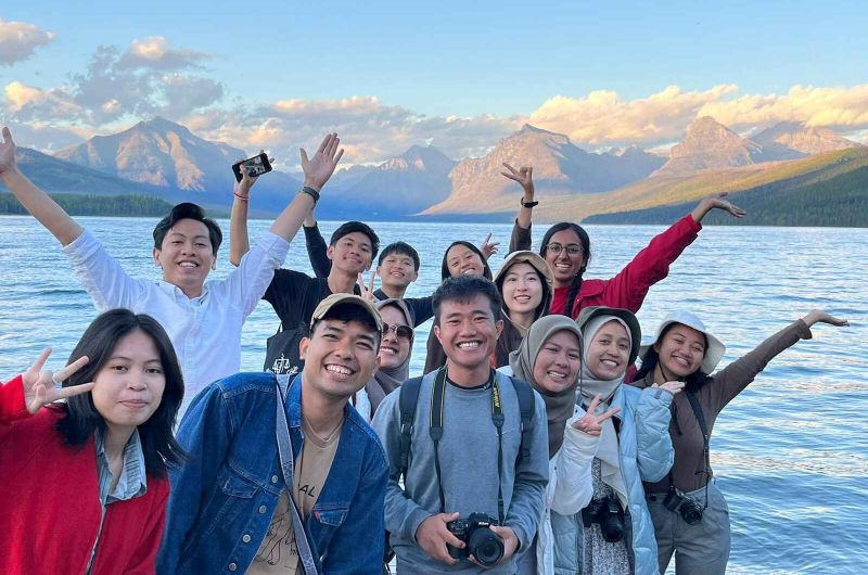 A group of diverse students posing joyfully by a lake with mountains in the background