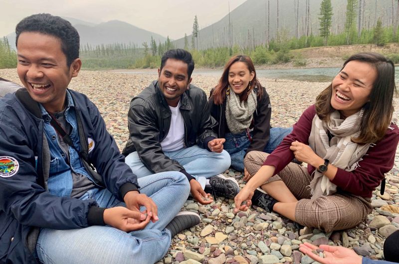 A group of four students laughing and sitting on a rocky riverbank