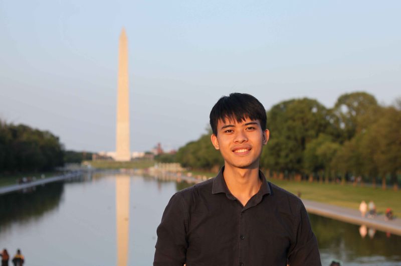 A young man smiling in front of the Washington Monument, reflecting in the nearby water at sunset