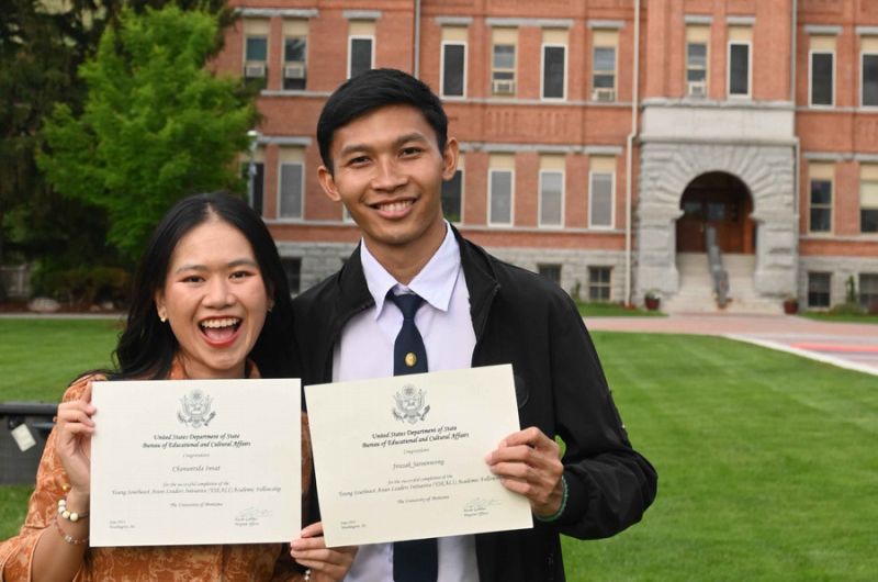 Two graduates proudly display their diploma certificates in front of a University Hall at UM