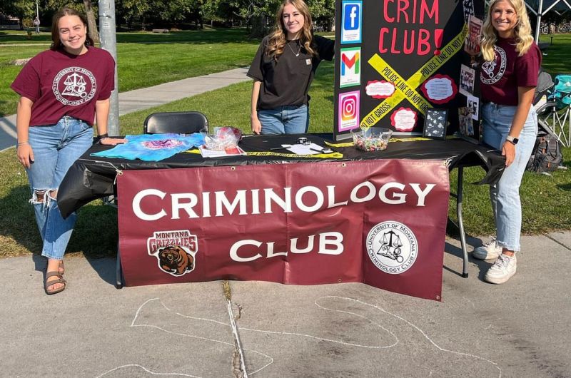 Crim Club leaders stand at an informational table with a chalk outline on the ground at the student Welcome event..