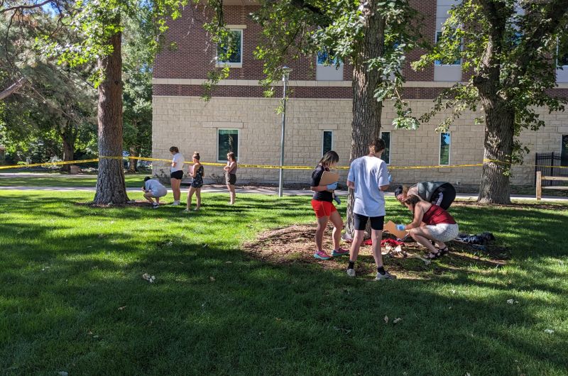 Groups of students outside surrounded by caution tape while investigating something on the ground.