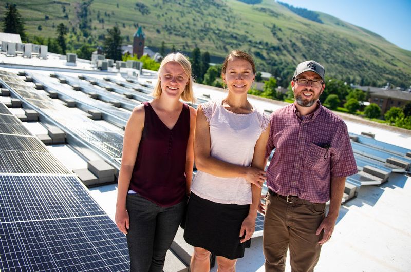 Eva, Maddie, and Paul smile on top of a sunny roof with solar panels in the background