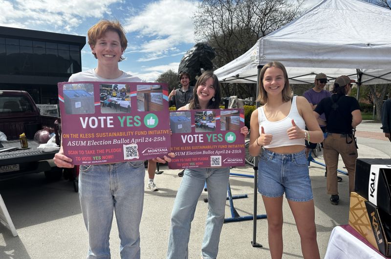 three students pose, two holding signs that share information about the Kless Fee increase campaign