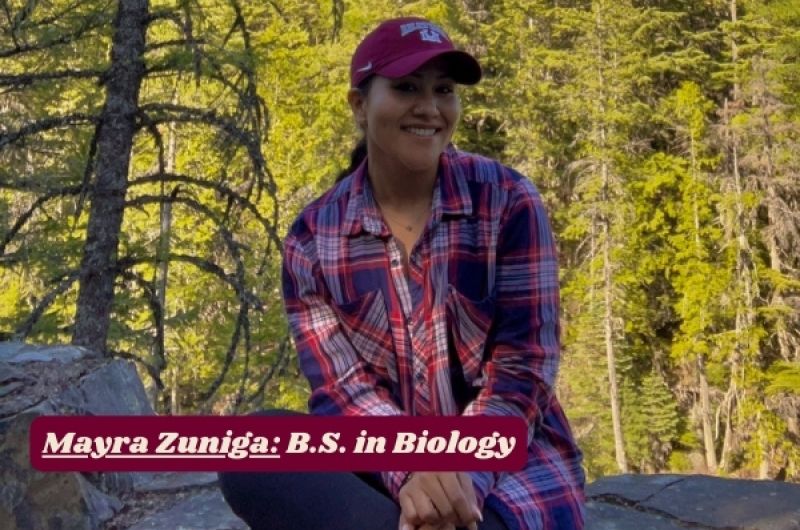 Mayra smiles while sitting on rock, dense forest in background