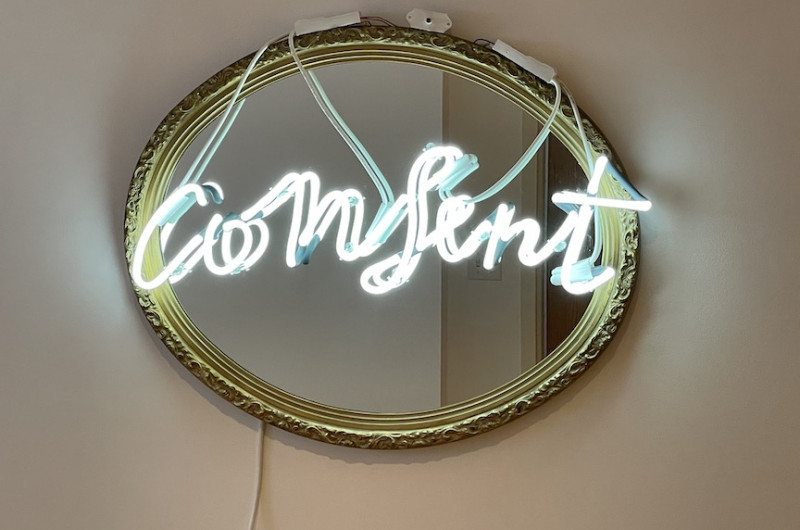 neon sign on mirror, neon is white and reads 'consent' in cursive, mirror has gold frame