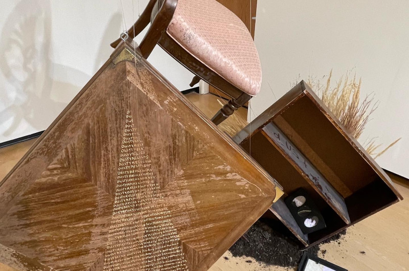 an end table with poem inscribed and a vintage chair with pink cushion hang suspended in the air next to a bookshelf that appears to be sinking into the ground