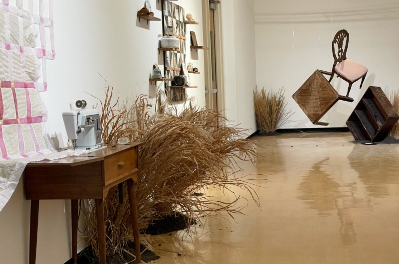 view of lefthand side of gallery including paper quilt, sewing machine, wood quilt, grasses, and hanging furniture in back